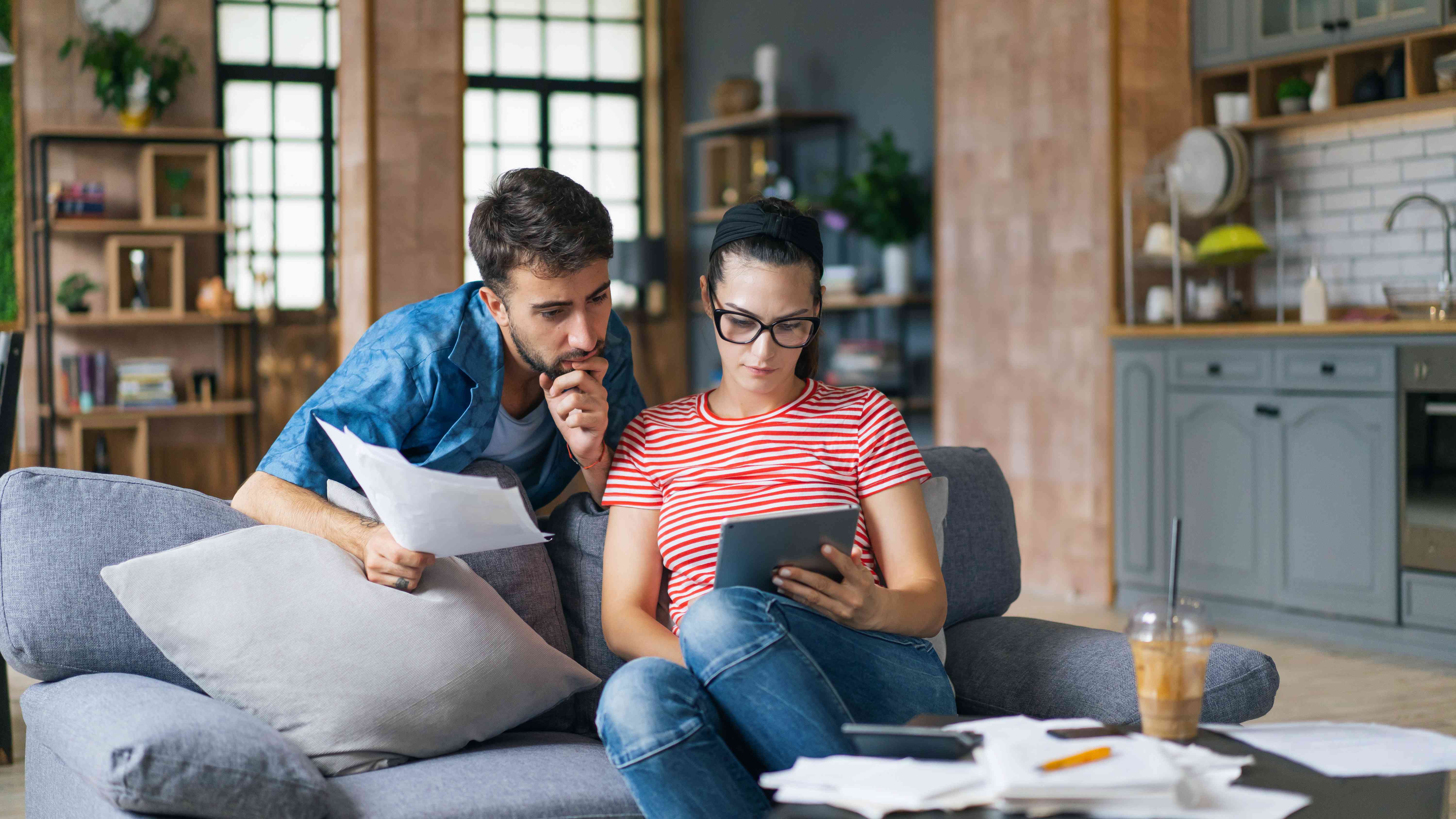 A couple sit together at a couch with a tablet computer and paperwork, analyzing their finances.