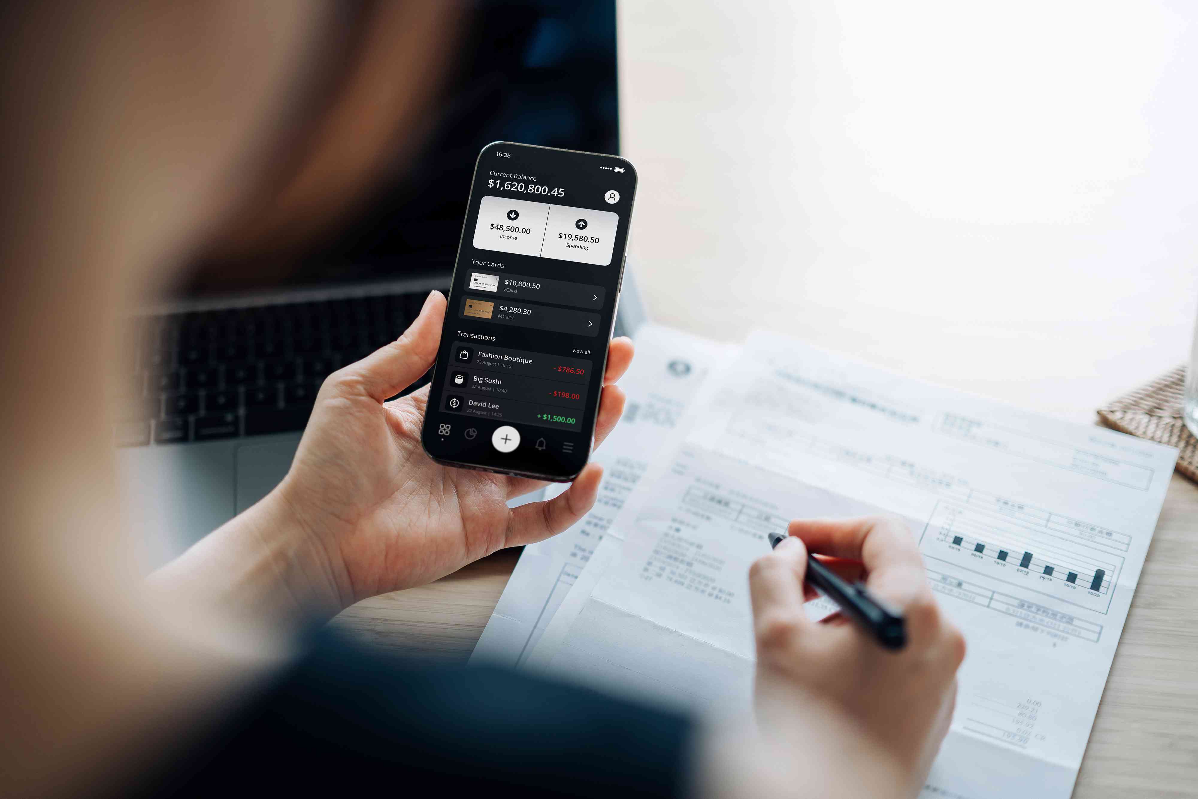 Over the shoulder view of young Asian woman checking online banking on smartphone while handling personal banking and finance at home